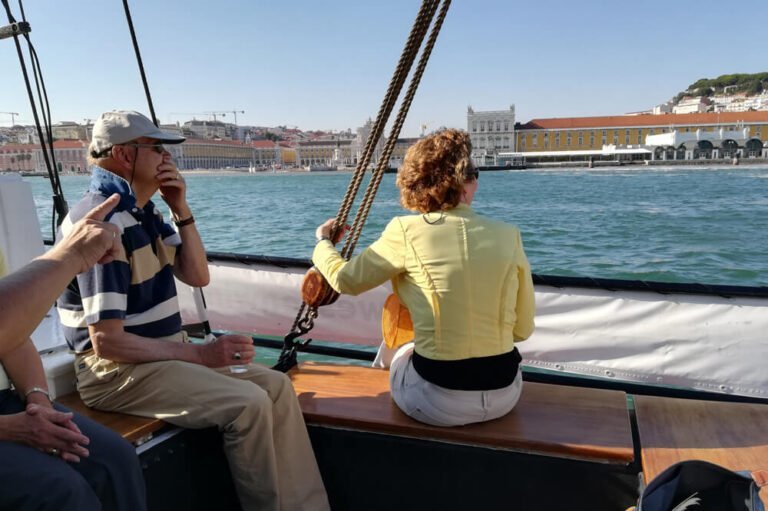 Pessoas a bordo de um veleiro clássico a ver a Praça do Comércio, a estacão Fluvial Sul e Sueste e o Castelo de S. jorge ao longe