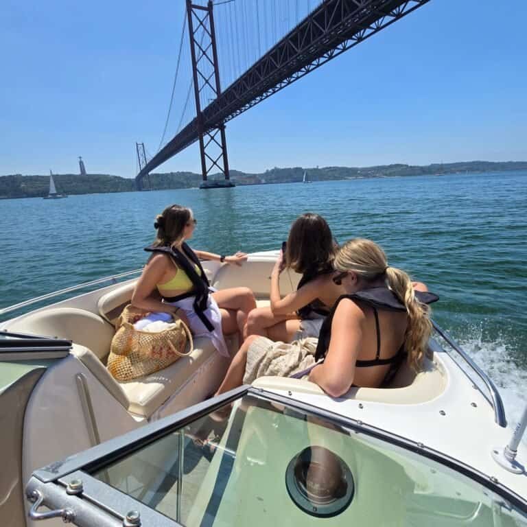 Grupo de amigas a bordo da lancha, navegando sob a Ponte 25 de Abril com vista para o Cristo Rei.